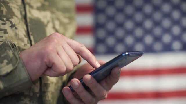 A soldier using a mobile phone in front of a U.S. flag.