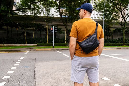 Man from behind wearing casual clothes and black sling bag stands at urban street crossing. Pedestrian waiting at red traffic light in city environment.