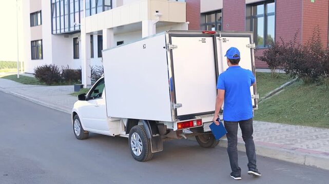 A male delivery man in a blue uniform opens a white delivery van and counts boxes of goods