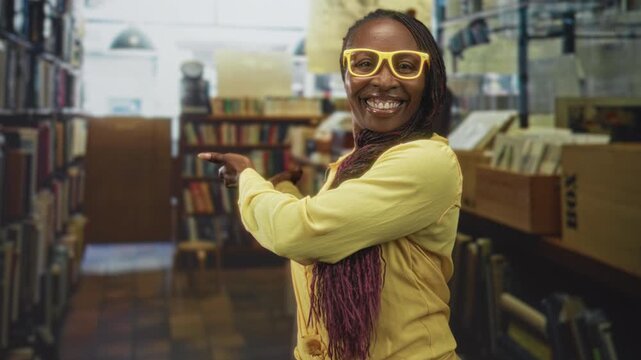 Young african american woman points finger at bookshelf while wearing yellow glasses in library; joyful discovery.