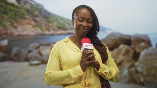 Woman holding a red microphone with news flag at chest on a rocky beach, yellow shirt, eyes closed and smiling while clasping the mic with both hands; serenity.