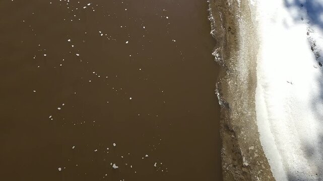 Top-down view of muddy spring runoff flowing along melting ice shelf in freshwater stream. Top-down view of sediment-laden freshwater stream flowing rapidly during spring snowmelt.