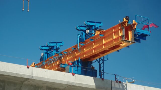 Handheld perspective looks up at the yellow and blue steel components of an industrial segment launcher resting on an elevated concrete transit guideway during the Surrey Langley SkyTrain construction