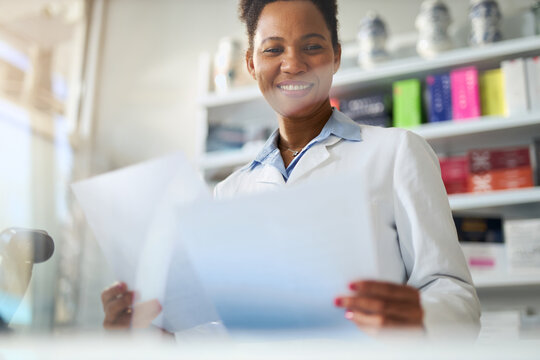Happy black female  pharmacist reading list of medicine while working in pharmacy