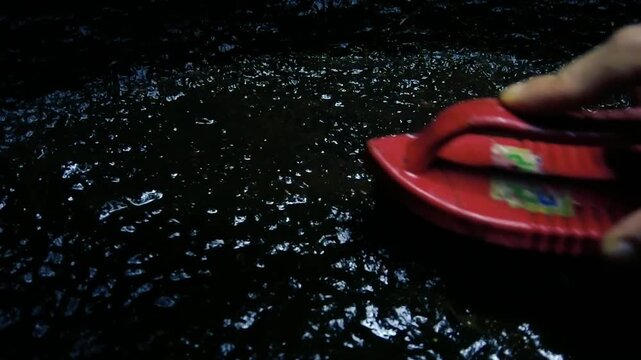 Hand scrubbing a dark wet mossy stone surface with a red plastic brush to clean dirt and algae
