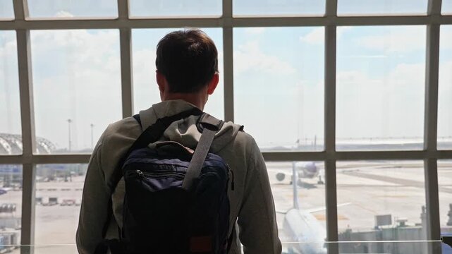 Traveler with backpack standing, looking through glass terminal window, modern airport terminal with airplanes on tarmac, travel vlog and aviation industry background, slow motion