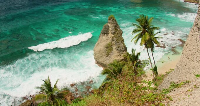 Sharp limestone peak rising from turquoise sea near sandy shoreline with palm trees in Nusa Penida Bali