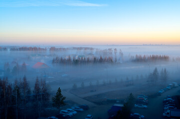 Obraz premium Spring morning view from a window: road, parking lot, trees and low fog at dawn. Cool atmosphere, soft light, misty mood. 