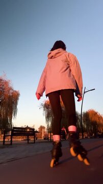Back view of young female rollerblader enjoys evening ride in urban park. Vertical stock video shows active recreation, smooth asphalt path, and bright sunset atmosphere