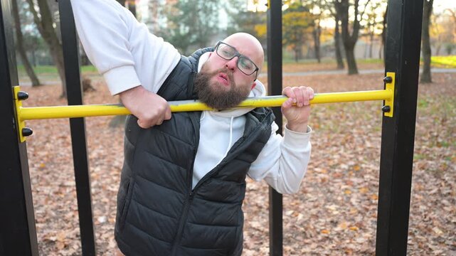 Bearded man performs pull-ups on yellow bar in serene park filled with fallen leaves, showcasing strength and determination while enjoying peaceful surroundings at sunset.