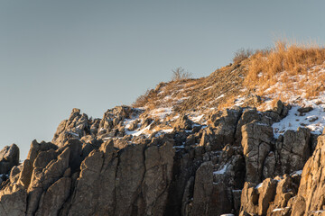 Rocky cliff landscape with snow patches and dry grass under clear sky during winter season, showcasing rugged terrain and natural beauty in a remote area © Denis Tuev
