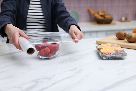 Woman wrapping bowl with fresh strawberries in cling film at light marble table, closeup