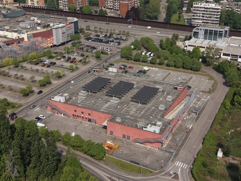 Aerial View of Commercial Building: An expansive commercial building complex, featuring a large parking lot and rooftop solar panels, is captured from an aerial perspective. Italy, Peschiera Borromeo