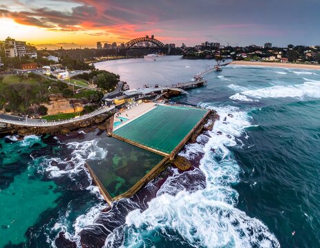 Aerial view captures a harbor with a bridge and a rock pool during a vibrant sunset, waves crashing, buildings in distance