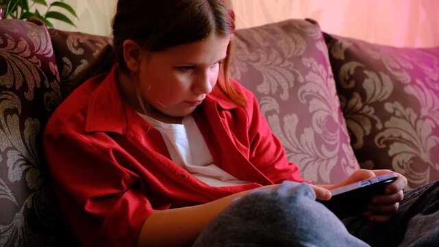 Teenager girl is engrossed in playing a game on their smartphone. They&rsquo;re sitting on a couch, fingers tapping the screen, completely absorbed in the virtual world. Their expression is focused
