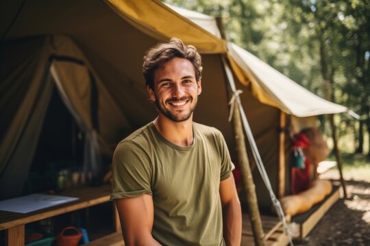 Smiling man with short hair wearing a green t-shirt stands in front of a camping tent in a lush forest, showcasing outdoor adventure and joyful experiences