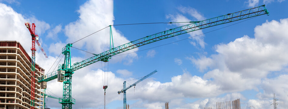 Two green tower cranes working at a residential construction site with workers in safety vests against a cloudy blue sky