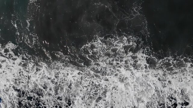 Top view of Atlantic waves reaching black stone shoreline, textured ocean surface and coastal water patterns