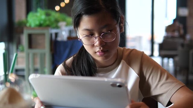 Young Asian student girl wearing glasses and earbuds using digital tablet for online learning and e-learning in a modern cafe atmosphere