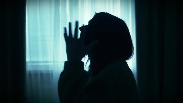 Young woman sitting alone in a dimly lit room, slowly raising hands in prayer near a window. Emotional scene of inner struggle, anxiety, loneliness and search for support, cinematic lighting and calm