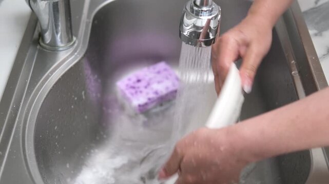 Washing a white plate in a kitchen sink using a sponge and soapy foam