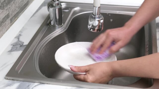 Washing a white plate in a kitchen sink using a sponge and soapy foam