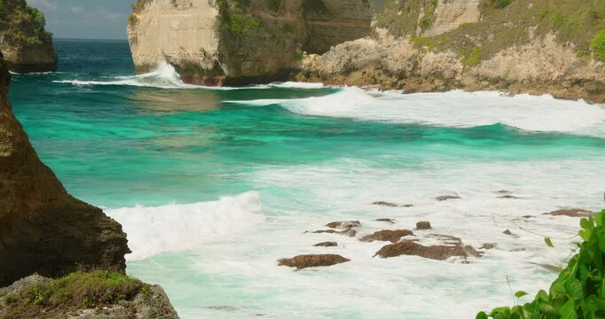 Foaming white waves break against dark coastal rocks in a tropical Indonesian cove
