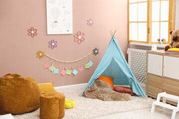 Colorful paper notes and beautiful decor hanging on beige wall near toy wigwam in stylish children's room with chest of drawers and poufs. Interior design © New Africa