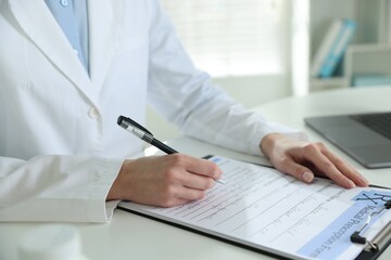 Fototapeta premium Doctor in medical coat writing on clipboard at white table in clinic, closeup