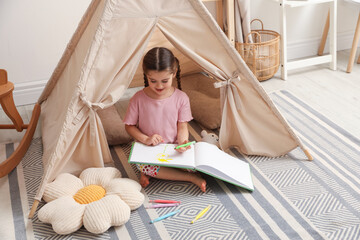 Little girl drawing in sketchbook near toy wigwam at home © New Africa