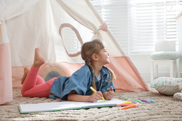 Little girl drawing in sketchbook near toy wigwam at home © New Africa