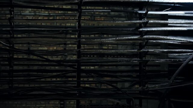 Dimly lit industrial basement featuring rusted metal cable trays, tangled wiring, and aged brick walls, illuminated by a single overhead lamp casts dramatic shadows and textures