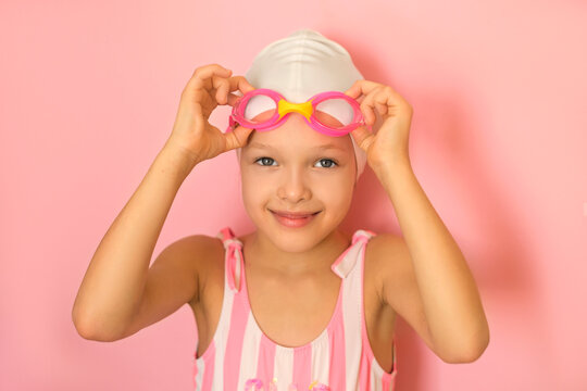 A pre-school athlete girl puts on swimming goggles. A child swimmer in a swimsuit on a pink studio background