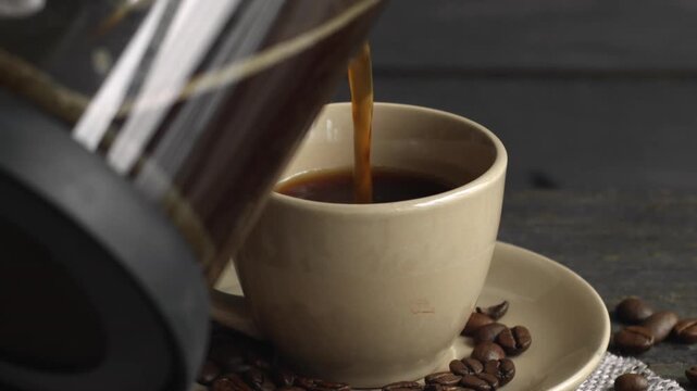 Pouring fresh hot coffee from a french press into a cup with roasted beans on a dark rustic wooden table