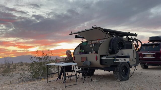 Mature woman dines beside her teardrop camper trailer during sunset in Anza Borrego Desert State Park. Beautiful landscape surrounds her.
