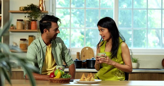Indian asian young husband and wife having coffee in modern kitchen platform with light hearted chit chat enjoying their morning beverage and talking together in a relaxed home environment
