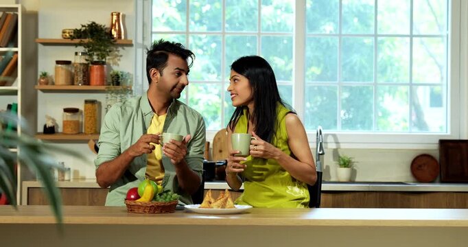 Indian asian young husband and wife having coffee in modern kitchen platform with light hearted chit chat enjoying their morning beverage and talking together in a relaxed home environment
