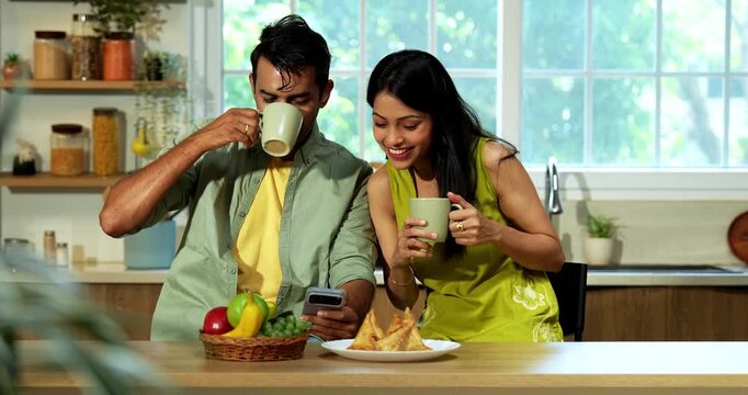 Indian asian young husband and wife having coffee at kitchen platform and surfing smartphone in joyful atmosphere in modern Indian home while sharing digital content and laughing during weekend