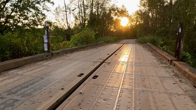 Low angle forward tracking of metal plated bridge with central gap and warm sheen reflecting sunset. At 0:08 dirt and gravel road curves right into dense forest with backlit foliage