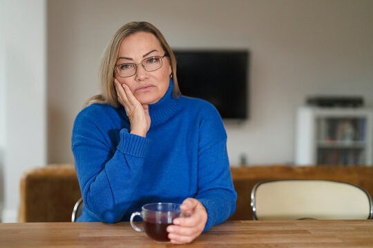 Woman sitting at kitchen table holding cup of tea and looking thoughtful in modern home