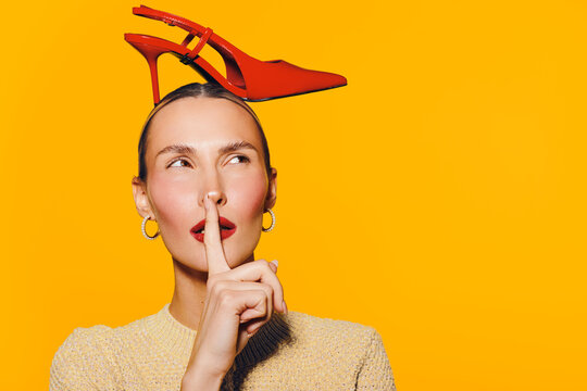 Young woman with red high heel shoe on head holds finger to lips in shushing gesture. Bright yellow background creates bold contrast. Concept of fashion, silence, and playful expression.