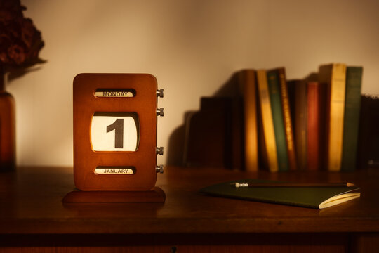 Vintage wooden perpetual calendar on a desk showing Monday January 1st. Retro office setup with old books, notebook, and pencil in warm, moody lighting. New Year's Day concept for business or study.