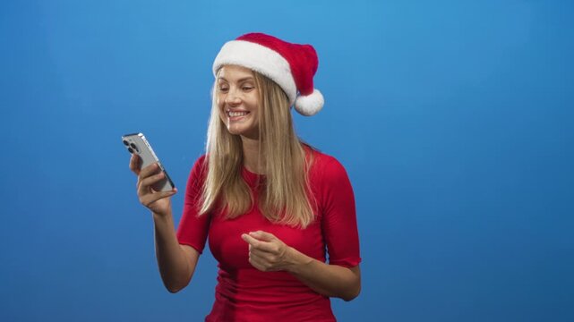 Blonde woman in a santa hat points at smartphone, grinning and gesturing with her hand while wearing a red top in a studio; holiday joy.