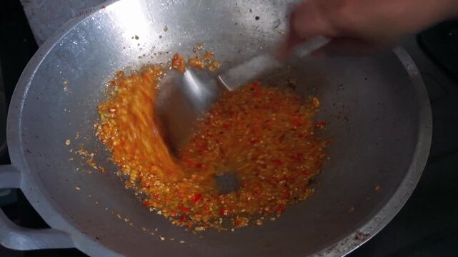 Hand of a chef stir-frying spicy red chili seasoning in a traditional metal wok with a spatula.