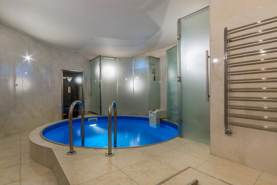 A round swimming pool is located in a modern spa area. Glass walls surround the pool, reflecting lights. The setting appears spacious and designed for relaxation in the evening