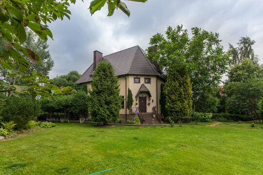 A house stands in a garden filled with greenery. Trees surround the home. The day is cloudy, and the neighborhood appears quiet. Grass is lush and neatly cut