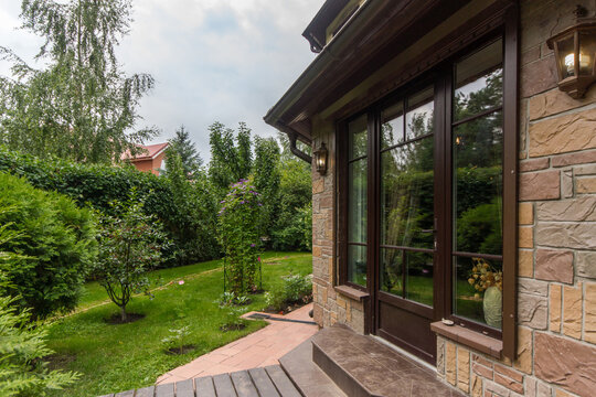 The scene shows a view from the house looking out at a green garden. The garden has trees and shrubs, and the sky is cloudy. The pathway leads deeper into the yard