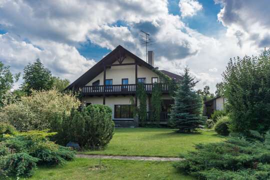 A house sits in a garden filled with green plants and trees under a cloudy sky. The area looks open and spacious, with no signs of people around