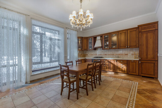 A kitchen features a large wooden table surrounded by chairs. Sunlight comes through big windows. Cabinets line the walls. The space is neat and tidy