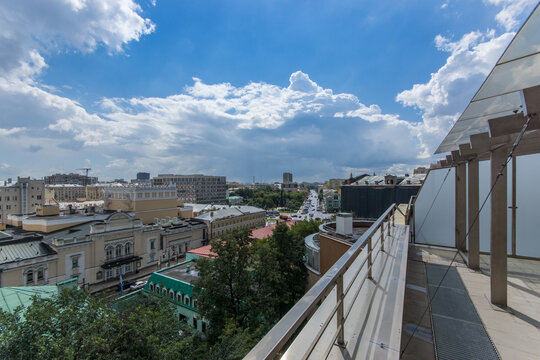 A balcony gives a wide view of the city filled with various buildings. The sky shows clouds and blue spaces, highlighting the daytime scene in a lively area